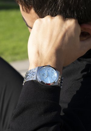 Montre en acier inoxydable argenté avec un cadran bleu clair. Les caractéristiques incluent des maillons métalliques, trois sous-cadrans et une accent rouge sur la lunette.