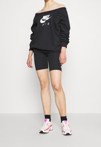 Černá mikina Nike bez ramenních popruhů s bílým logem, v kombinaci s černými cyklistickými šortkami a bílými teniskami s růžovými a oranžovými akcenty.