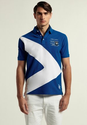 Camisa polo azul con un diseño diagonal blanco, hecha de algodón, con un cuello y detalles de logo bordados. Acompañada de pantalones de color claro.