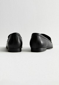 Mocasines de cuero negro con textura suave, tacón bajo y diseño minimalista. Presentan una puntera redondeada y detalles de costura sutiles.