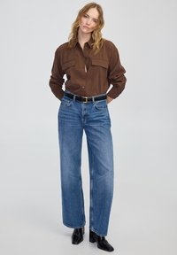 Brązowa koszula zapinana na guziki z dwoma kieszeniami na piersi, zestawiona z wysokim stanem niebieskimi spodniami w szerokim kroju oraz czarnymi botkami na obcasie.