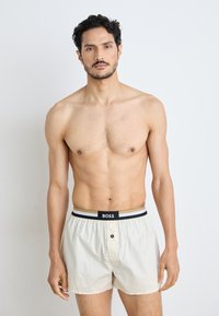 Crème-kleurige boxers gemaakt van lichte stof, met een zwarte tailleband met een logo en horizontale accentstrepen. Drukknoopsluiting aan de voorkant.