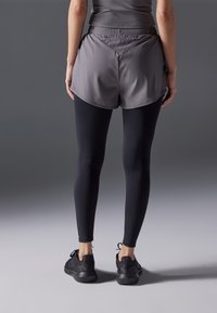 Urheilushortsit harmaana sivuhalkioilla mustien legginsien päällä, yhdistettynä mustiin urheilukenkiin. Varustettu joustavalla vyötäröllä ja sileällä pinnalla.