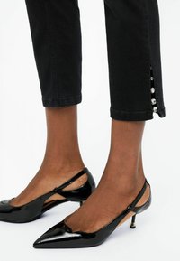 Pantalons noirs cropped avec des boutons décoratifs sur le côté ; associés à des escarpins noirs brillants à talons ouverts avec une pointe et un petit talon.