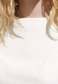 Haut blanc à manches courtes avec un design simple, fabriqué dans un tissu lisse. Décolleté dégagé. Cheveux doux et fluides partiellement visibles.