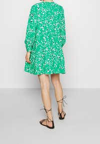 Vestido floral verde con mangas largas y diseño en capas, que presenta patrones de hojas blancas. Se combina con sandalias negras de tiras.