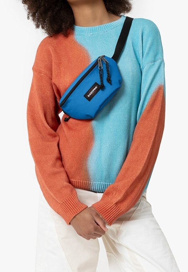 Personne portant un pull bleu et orange avec un sac bandoulière bleu Eastpak et un pantalon blanc, mains jointes devant.