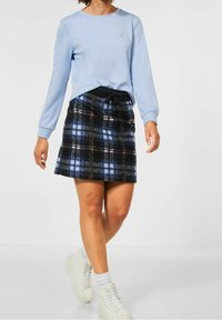 Sweatshirt bleu clair à manches longues et col rond, associé à une mini-jupe à carreaux noirs et bleus et des baskets blanches.