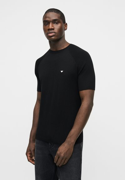 Νέος άντρας που φοράει στενή μαύρη κοντομάνικη μπλούζα με μικρό λευκό λογότυπο στο στήθος, συνδυασμένη με σκούρο τζιν, μπροστά σε ένα απλό φόντο.