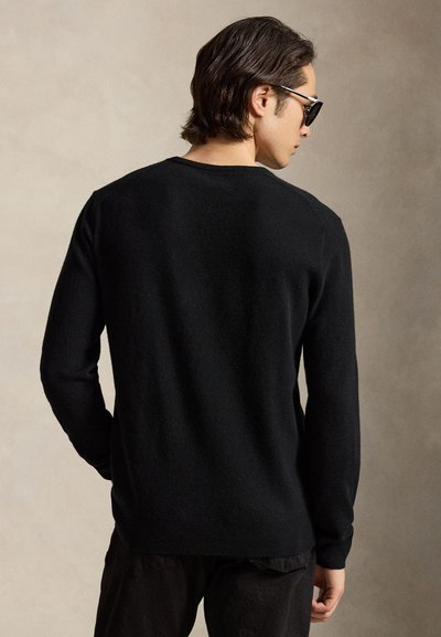 Mann mit dunklem Haar, der einen schwarzen Pullover und Sonnenbrille trägt, steht mit dem Rücken zu einem schlichten hellen Hintergrund.
