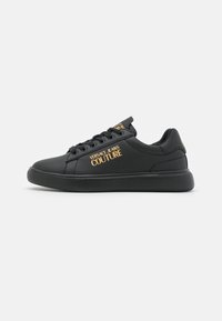 Svart sneaker med slät syntetisk material, rund tå, snörning och guld "Versace Jeans Couture"-logotyp på sidan.