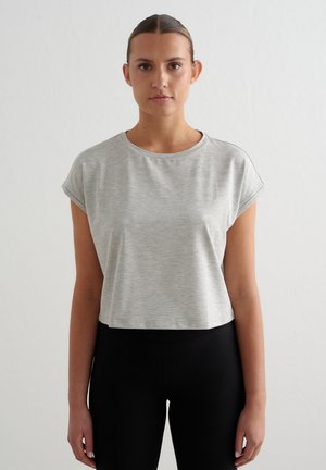 Ung kvinde med håret i en knold iført en lysegrå kortærmet crop top og sorte høj taljede leggings, står mod en ensfarvet baggrund.