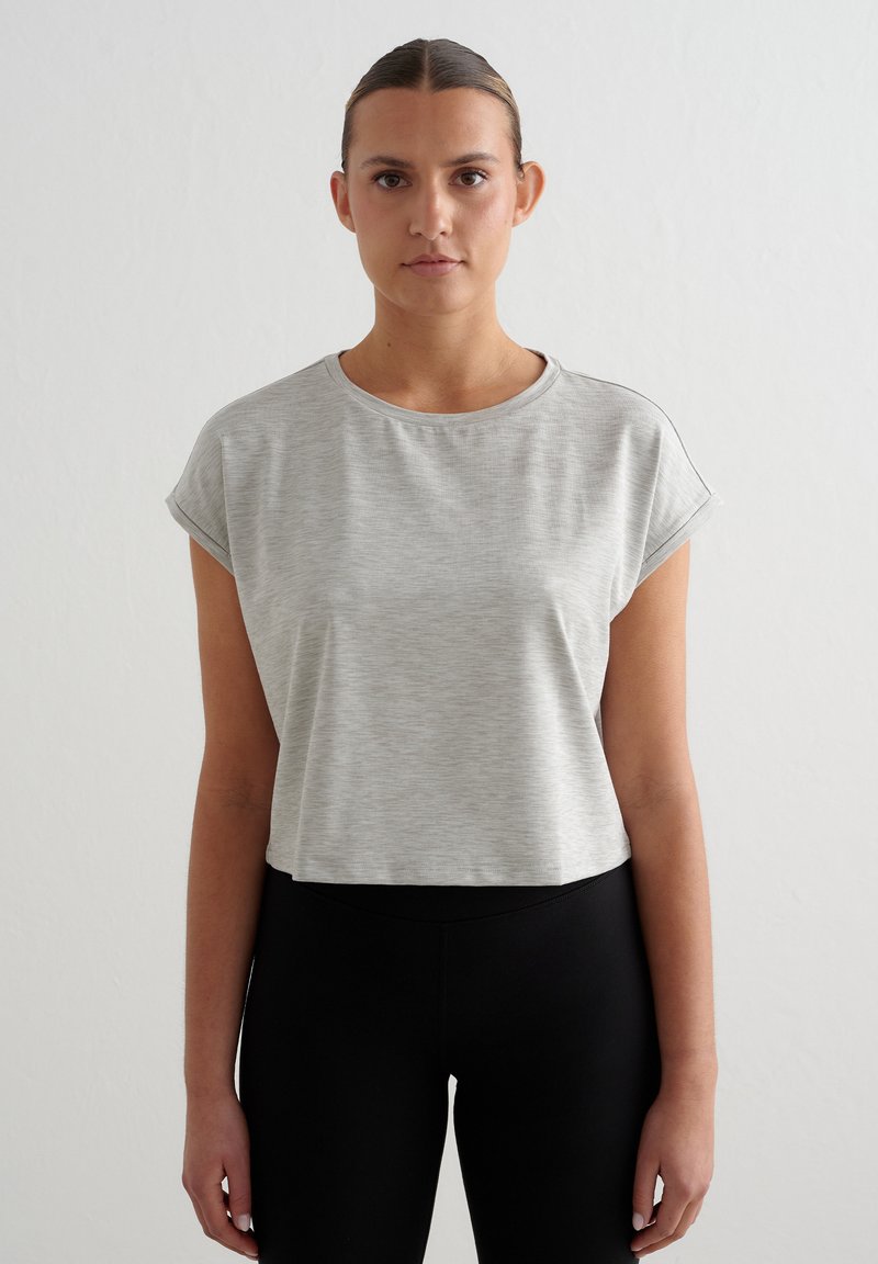 Jeune femme avec les cheveux en chignon, portant un crop top gris clair à manches courtes et des leggings noirs taille haute, debout sur un fond uni.