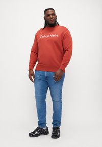 Červená mikina s bielym logom "Calvin Klein", skombinovaná s modrými džínsami a čiernymi teniskami. Úzky strih, hladká textúra, dizajn s okrúhlym výstrihom.