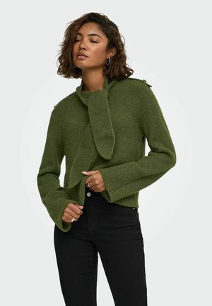 Femme en pull vert tricoté avec écharpe attachée et pantalon noir, debout devant un fond clair uni.