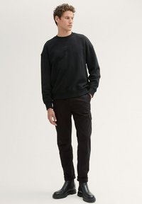 Svart sweatshirt med ribbade muddar och fåll, med en liten präglad logotyp på bröstet. Kombinerad med svarta cargobyxor och ankelstövlar.