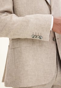 Beżowa lniana marynarka o slim fit, subtelnej strukturze i pięciu guzikach na mankietach. Szczegóły szwów oraz jedna kieszeń na piersi.