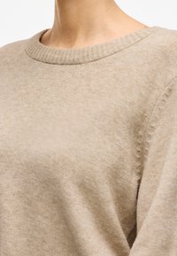 Beżowy dzianinowy sweter z prążkowanym dekoltem, charakteryzujący się miękką fakturą i subtelnym detalem przeszycia na rękawie.