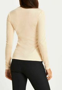 Langärmliges Oberteil in Hellbeige, aus einem glatten, elastischen Stoff gefertigt. Figurbetontes Design mit rundem Ausschnitt und minimalen Nähten. Kombiniert mit schwarzen Leggings.
