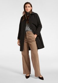 Zwarte waterdichte jas met knopen, grijze coltrui, beige wijde broekspijpen en bruine puntige platte schoenen. Strakke lijnen en minimalistisch design.