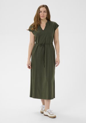 Kobieta ubrana w oliwkowo zieloną, midi sukienkę z krótkim rękawem i wiązaniem w talii oraz białe trampki, stojąca na tle jednolitego, jasnego tła.