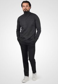 Jersey de cuello alto de color carbón, hecho de una suave tela de punto, combinado con pantalones negros texturizados y zapatillas blancas. El modelo está de pie de manera casual con las manos en los bolsillos.