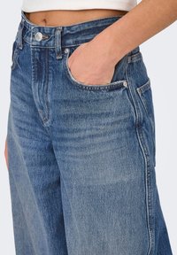 Jeans in denim blu a gamba ampia con vita alta, dotati di chiusura con bottone, tasche frontali e un lavaggio sbiadito per aggiungere texture.