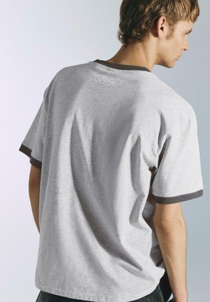 T-shirt en coton gris avec une coupe décontractée, présentant une bordure foncée autour des manches et un logo "Ford" subtil brodé dans le dos.