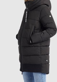 Chaqueta de invierno negra acolchada con capucha, cierre frontal, bolsillos laterales y dobladillo acanalado, llevada sobre unos jeans de color azul claro.