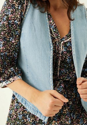Frau richtet eine hellblaue Jeansweste über einem schwarzen Blumenkleid mit rosa, blauen und grünen Blumen, die Ärmel bis zu den Ellbogen hochgekrempelt.