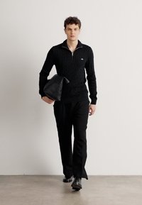 Black cable-knit sweater with zip collar, paired with black trousers. Model holds a black leather bag while walking on a light concrete surface.