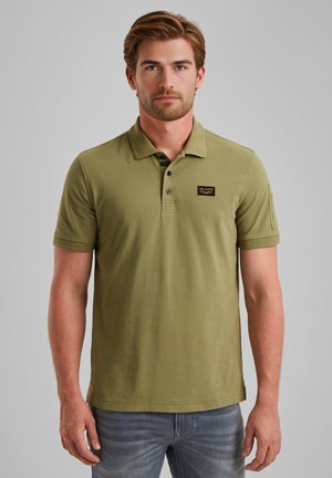Olijfgroene polo met korte mouwen van zacht katoen, voorzien van een zwart logo-embleem en een drieknoopsluiting, gecombineerd met lichtblauwe jeans.