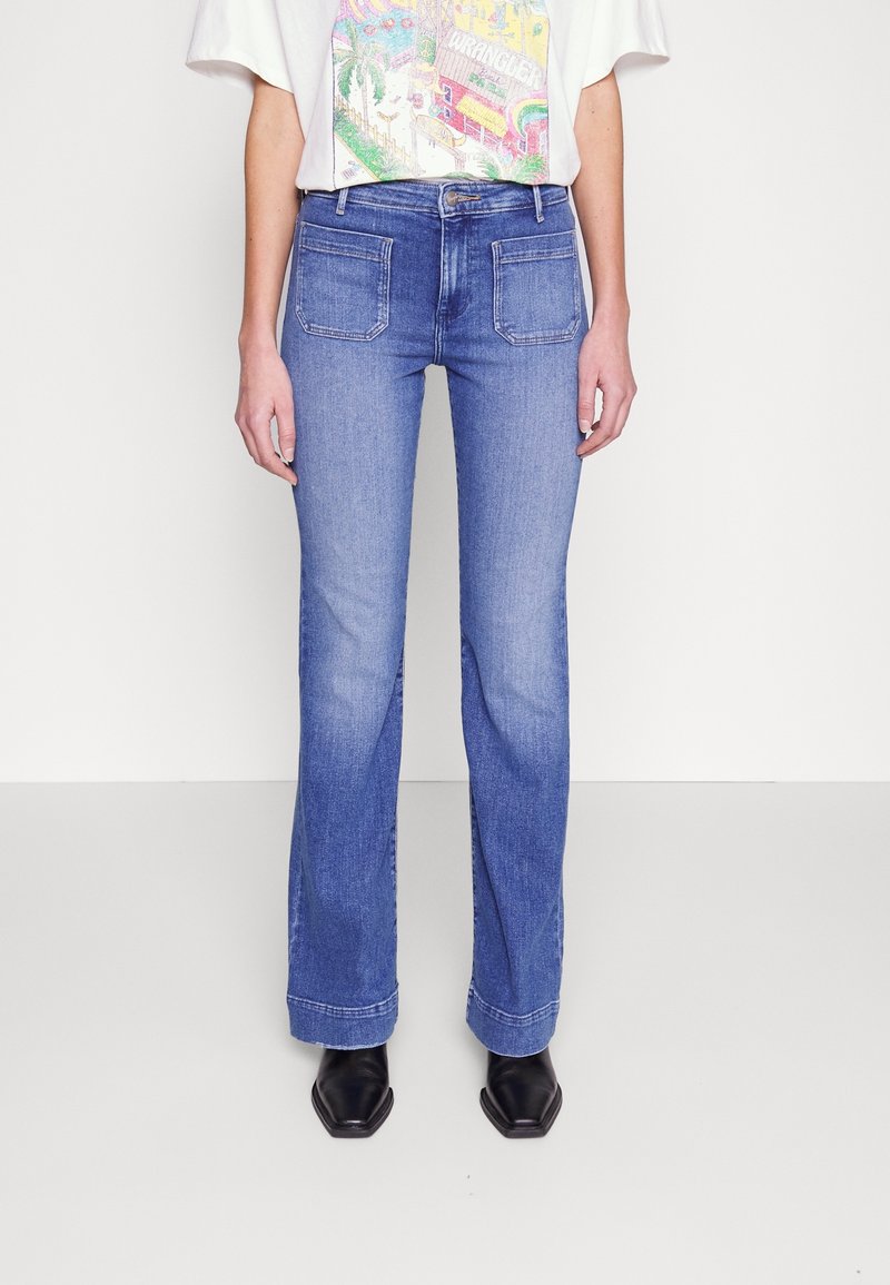 Femme portant un jean bleu droit avec des poches plaquées à l’avant, un t-shirt blanc graphique rentré et des chaussures noires à bout pointu.