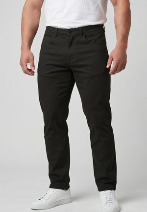 Man draagt strakke zwarte broek, witte sneakers en een wit t-shirt en staat tegen een effen achtergrond.