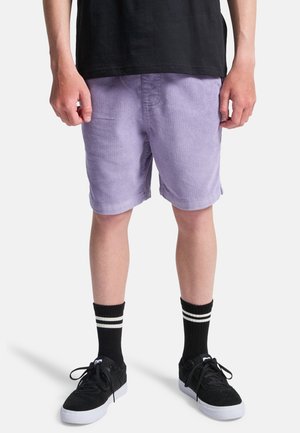 Λιλά βερμούδα από κοτλέ με ίσια γραμμή και γυριστά τελειώματα. Φοριέται με μαύρα αθλητικά παπούτσια και μαύρες κάλτσες με λευκές ρίγες.