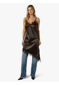 Sieviete, kas valkā melnu satīna kleitu ar mežģīņu maliņu, slānis pāri zilajām džinsu biksēm un melnām kurpēm ar smailiem purngaliem, stāvot pret baltu fonu.