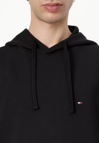 Zwarte hoodie van zachte stof, met een trekkoordcapuchon en een klein geborduurd logo in rood, wit en blauw op de borst.