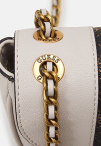 Lys beige taske med en tekstureret overflade, der har gyldent hardware og en kædestrop. Bemærkelsesværdige mærkeaccenter i runde metalnitter.