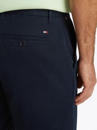 Calças de algodão azul-marinho com um bolso traseiro de botão e um pequeno emblema em vermelho, branco e azul. Textura suave e corte ajustado.