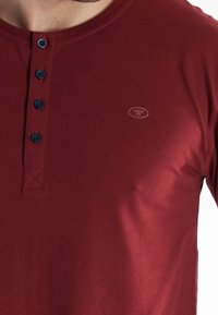 Bordeaux lange mouw shirt met een ronde hals, voorzien van een knoopsluiting, contrasterende marineblauwe knopen en een klein logo op de borst.