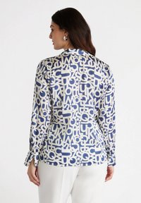 Long-sleeve blouse in a cream fabric with a bold blue abstract pattern, featuring a classic collar and button cuffs. Smooth texture.