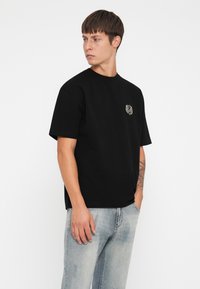 Zwart katoenen T-shirt met korte mouwen en een ronde halslijn, met een klein gouden embleem op de borst. Gestyld met lichtblauwe jeans.