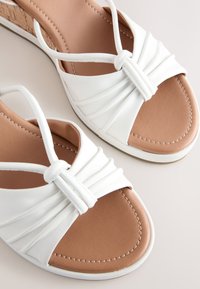 Sandales blanches avec un design torsadé à l'avant, semelle intérieure marron coussinée et talon texturé effet liège. Tige en cuir lisse avec détails cousus.