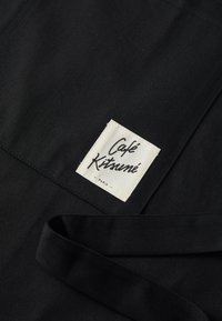 Svart bomullsforkle med en strukturert finish, med en hvit etikett sydd med "Café Kitsuné Paris" i svart. Justerbare bånd.