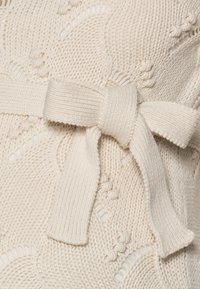Kremowy sweter z dzianiny, z teksturowanym wzorem i pasującym paskiem zawiązanym w talii, z prążkowanymi akcentami wzdłuż brzegów.