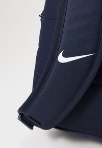 Mochila azul-marinho com tecido texturizado, apresentando um logótipo branco da Nike na alça e bolsos laterais discretos com fecho éclair.