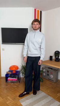 Felpa grigia chiara con zip e logo ricamato, abbinata a pantaloni di denim scuri a gamba larga e scarpe bordeaux lucide, in un ambiente moderno.