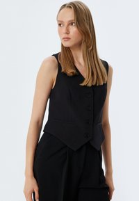 Schwarzes ärmelloses Hemd mit V-Ausschnitt und Fünf-Knopf-Verschluss. Verfügt über zwei seitliche Taschen und eine taillierte, leicht ausgestellte Silhouette.