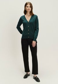 Taehlfarbener Cardigan mit V-Ausschnitt, Knopfschluss und gerippten Manschetten, kombiniert mit schwarzen, taillierten Hosen und Slippern, vor einem hellen Hintergrund präsentiert.