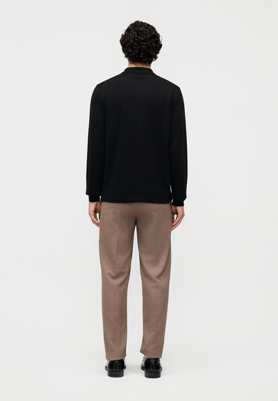 Άνδρας με σγουρά μαλλιά, φορώντας μαύρη μακρυμάνικη μπλούζα, μπεζ παντελόνι και μαύρα παπούτσια, στέκεται με την πλάτη προς τα εμπρός μπροστά σε απλό φόντο.